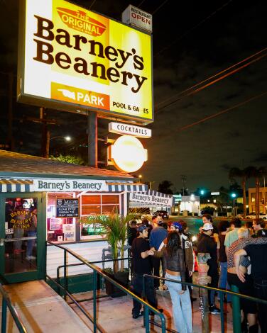 A crowd stands outside under a big yellow lighted Barney's Beanery sign at the restaurant
