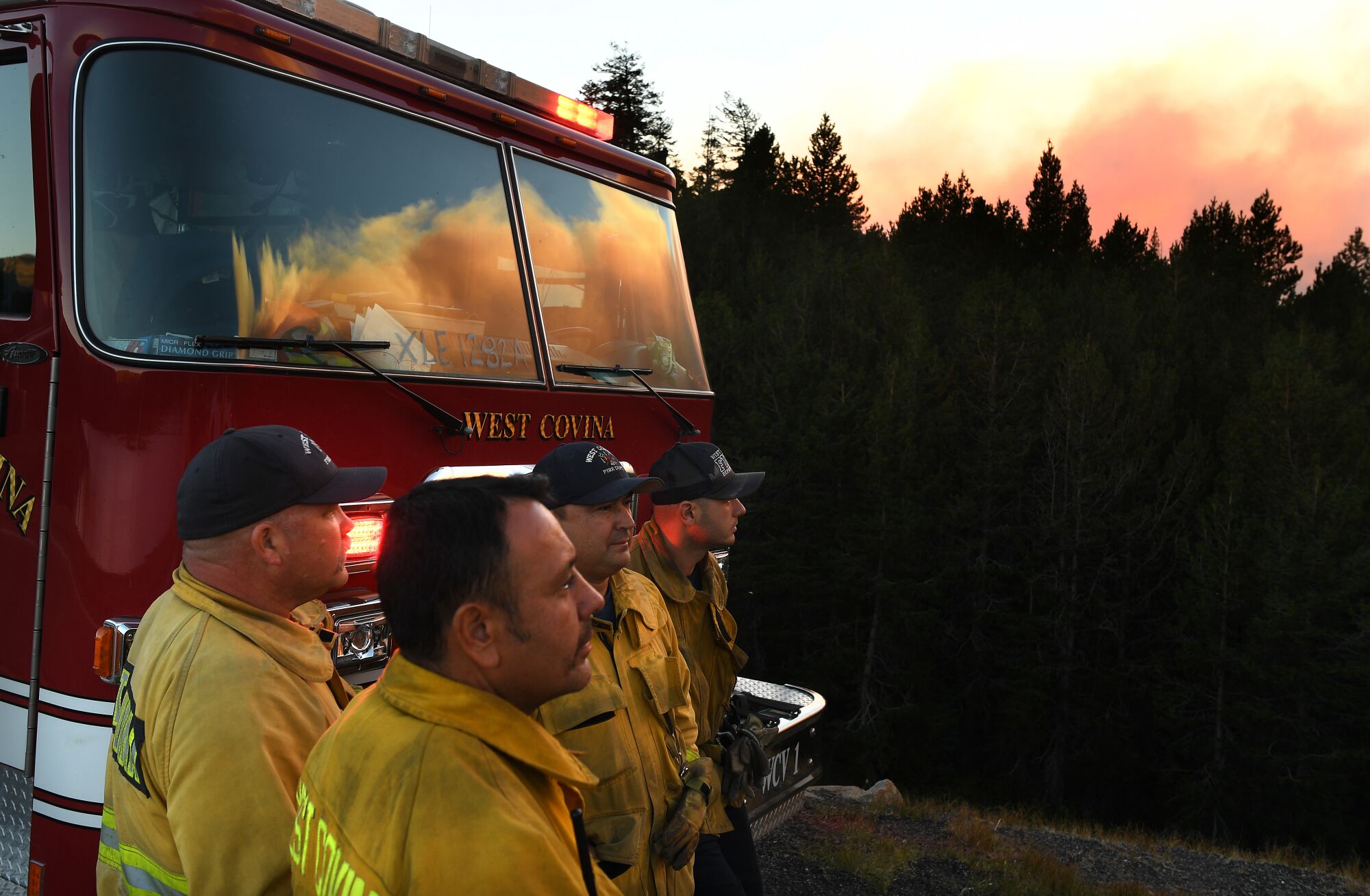 Firefighters stand in front of a firetruck and look toward a smoky sky.