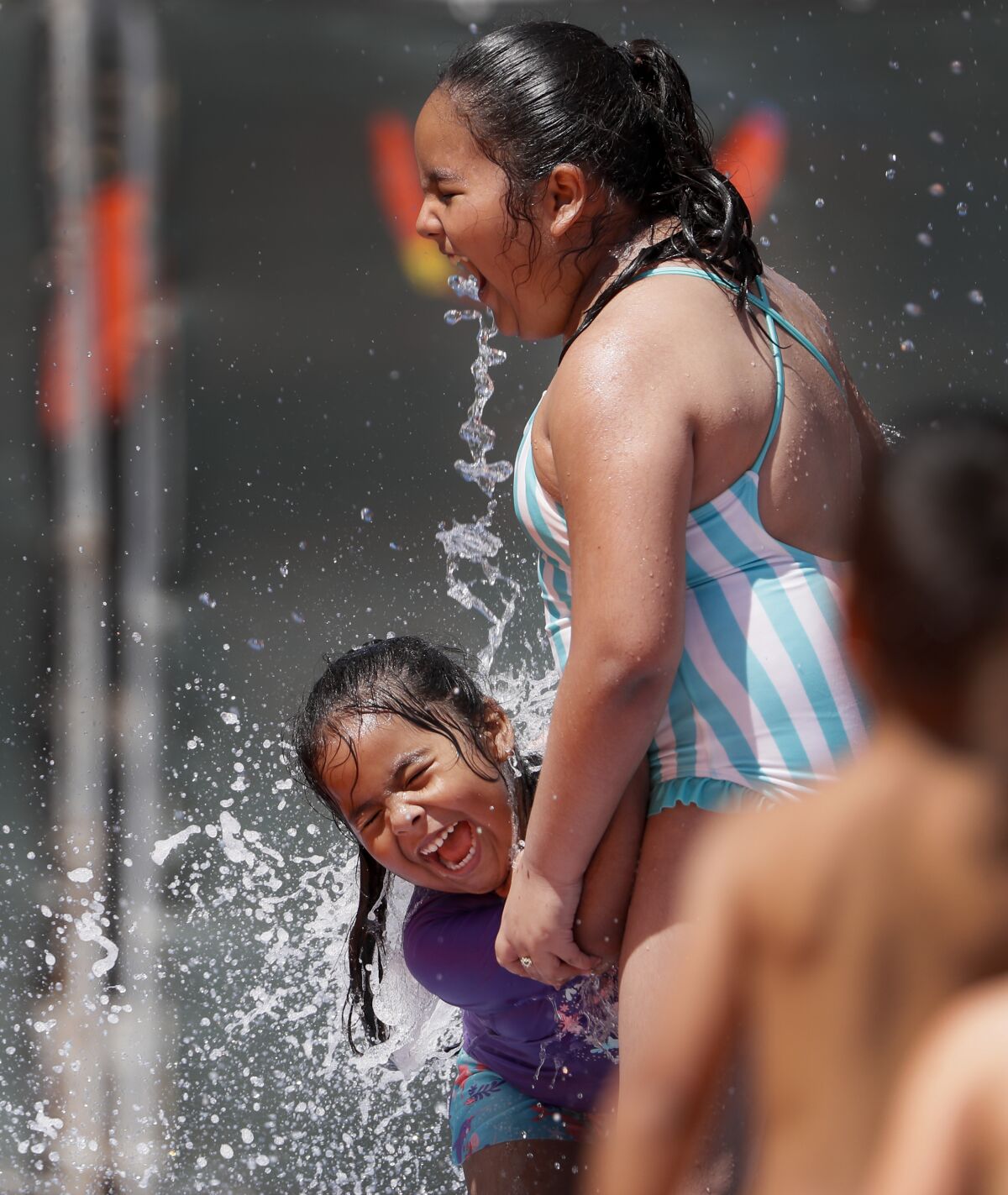 Two young girls laugh as they stand in a spray of water at a play area