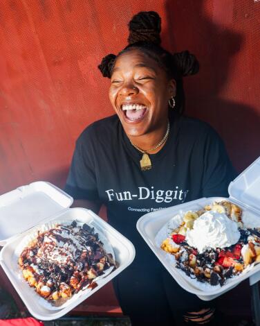 Cheyenne Brown laughs while holding two styrofoam containers filled with funnel cakes covered with toppings.