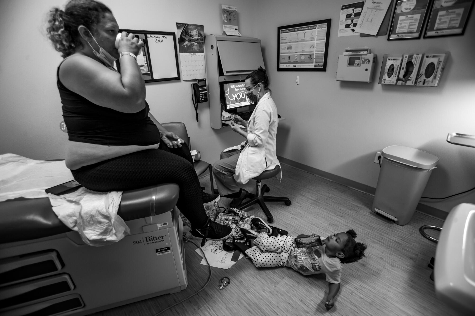 A woman sitting on an exam table drinks from a cup as a doctor sits nearby and a child lies on the floor