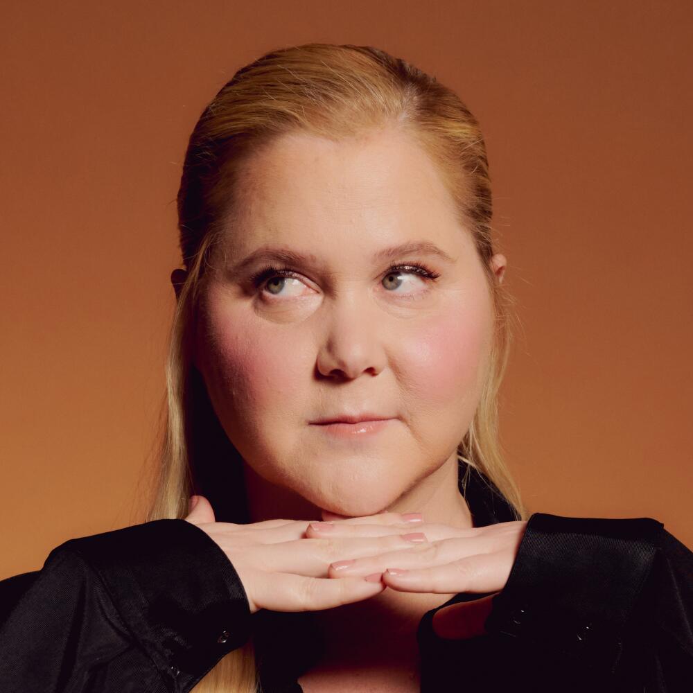 A close portrait of Amy Schumer who holds her hands below her chin and looks to the side.