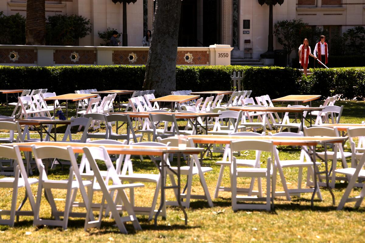 Rows of vacant chairs and tables at USC as two graduates with cardinal and gold sashes look on from the background