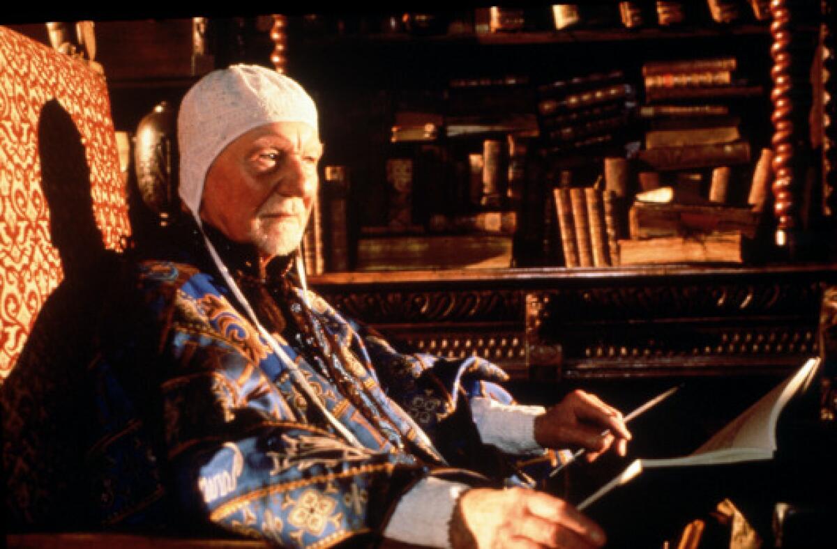 An older man wearing a cap and shawl sits in a chair surrounded by books.