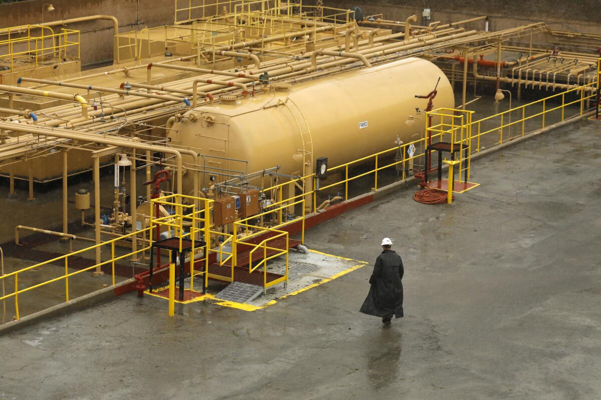 An inspector in hard hat and black coat at an oil facility with tanks and pipes