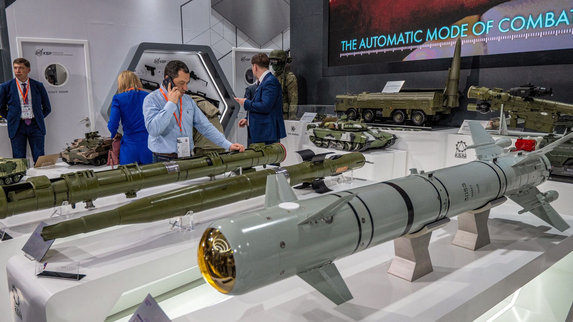 A man speaks on his cellphone as he stands near one of a few projectile-like weapons on display. Behind him are other people 