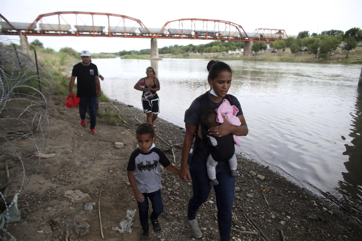 A woman carrying an infant holds the hand of a young child, while two people walk behind them along a river 