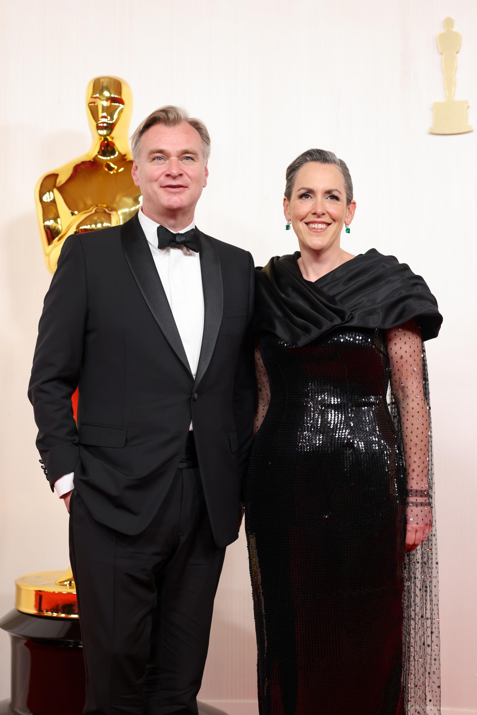 Christopher Nolan and Emma Thomas, in black, pose in front of an Oscar statue. 