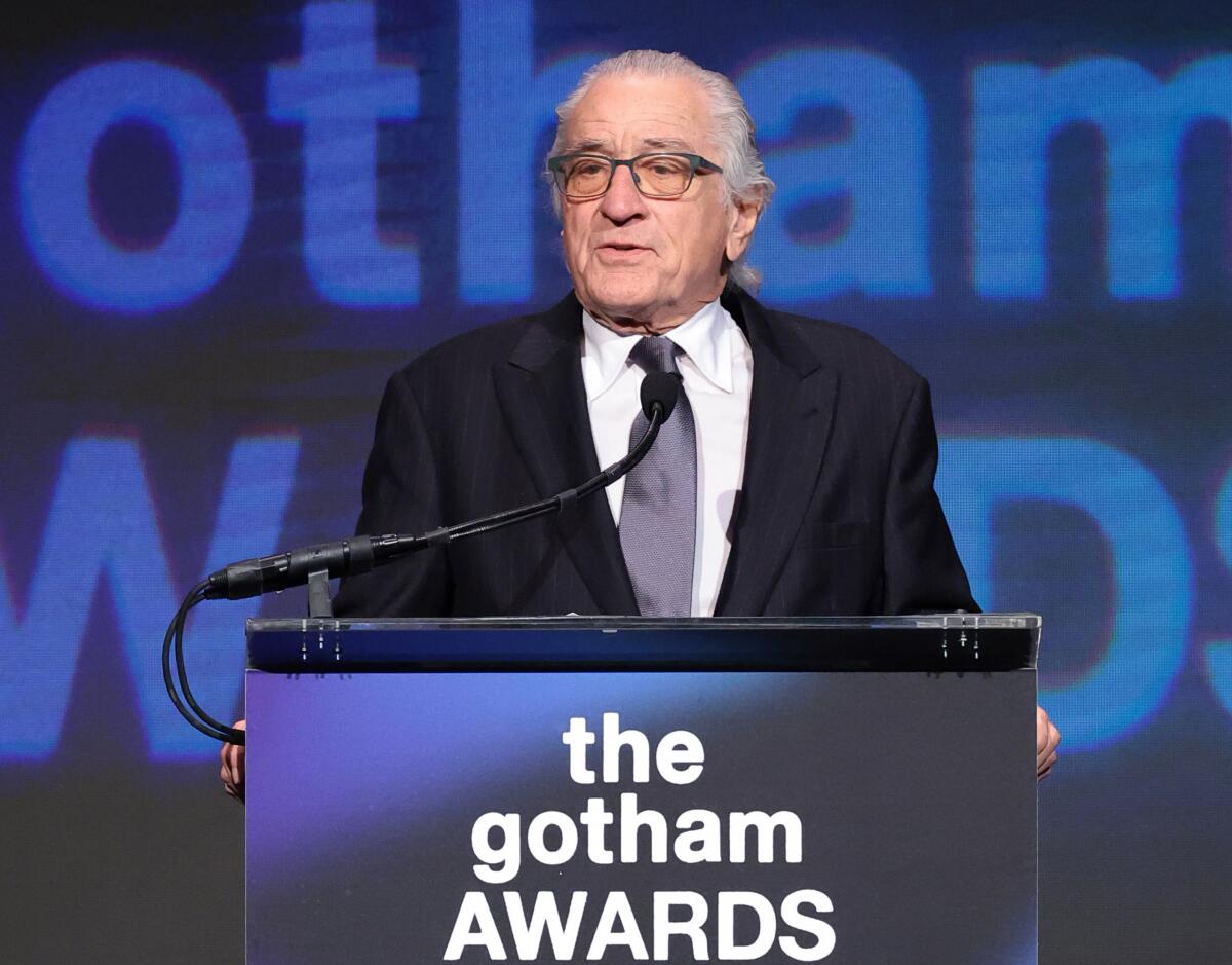 Robert De Niro speaks from behind a lectern while wearing a dark suit and lighter tie at the 2023 Gotham Awards