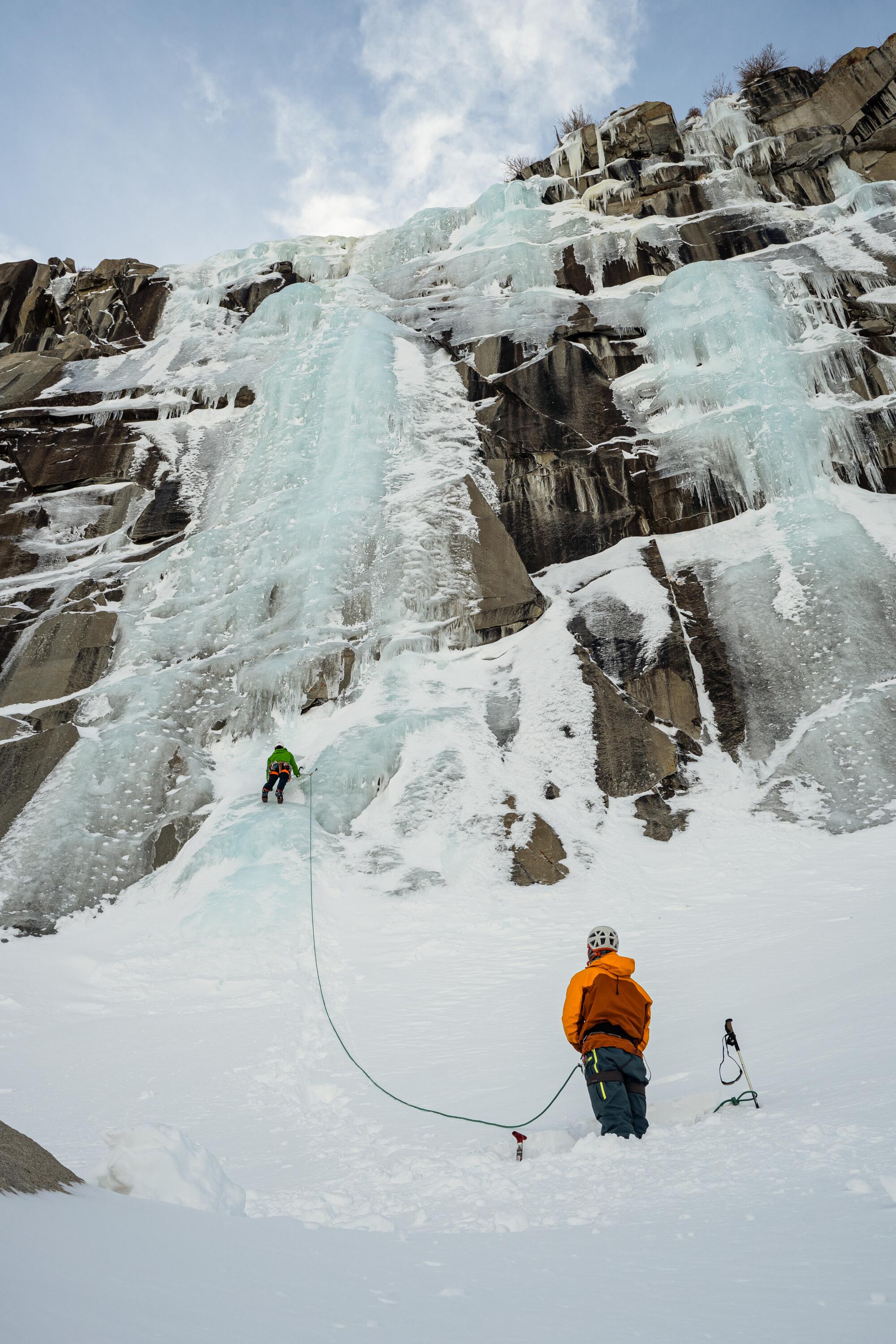 An ice climber stands at the base of a frozen waterfall, watching another climber some distance ahead