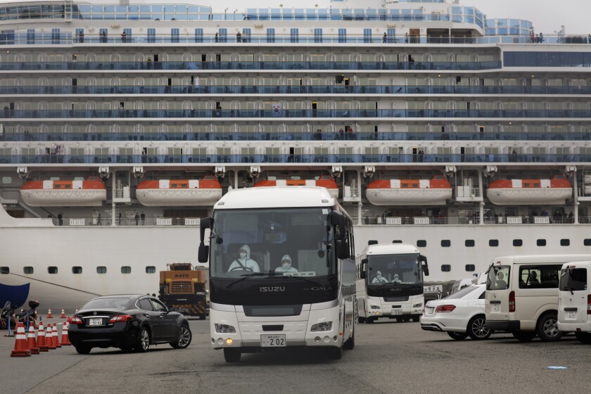 A lot with a bus driven by someone in medical protective gear and other vehicles in front of a docked cruise ship.