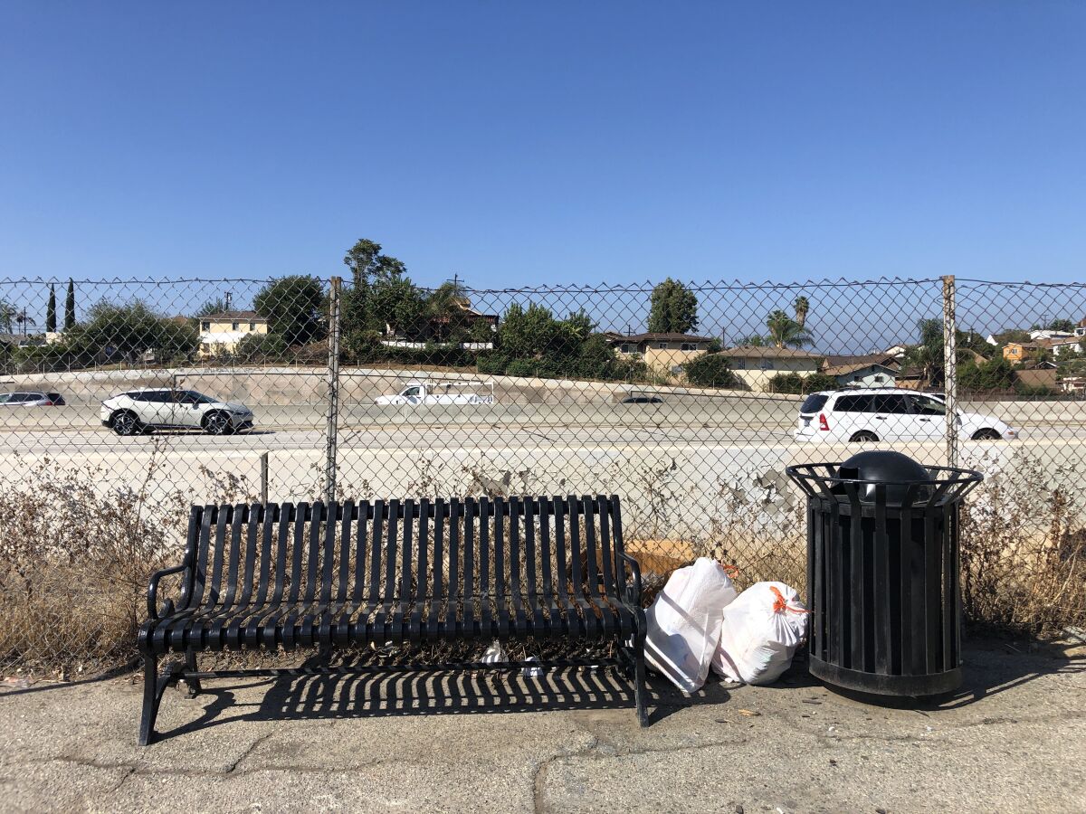A black metal bench and a garbage can sit before a chain link fence and a busy freeway
