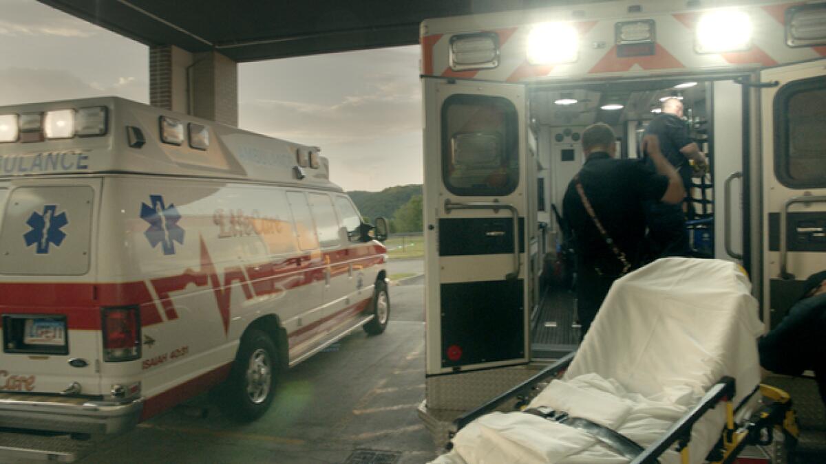 Two paramedics in the back of an ambulance, adjacent to an empty stretcher, with another ambulance parked alongside.