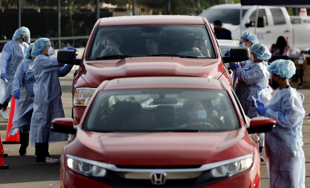 Masked and gowned healthcare workers surround cars at a COVID test site.