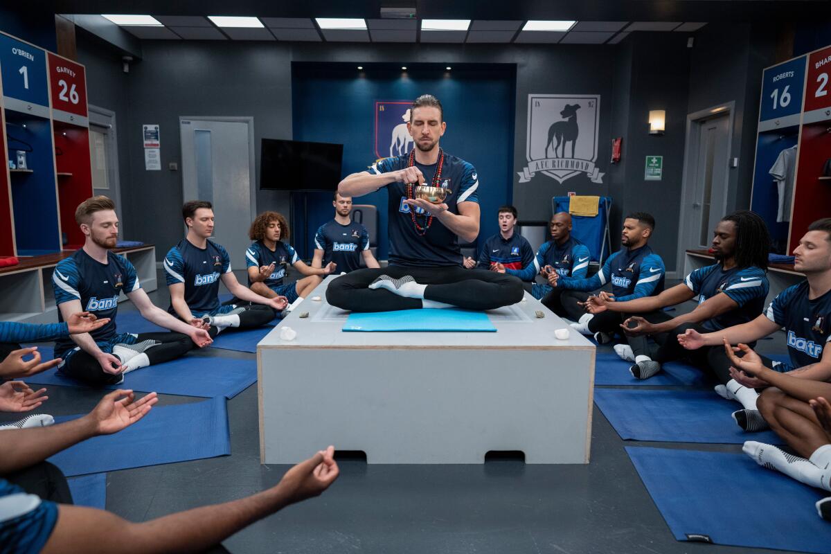 A man meditating on a pedestal in a locker room, surrounded by a group of men meditating