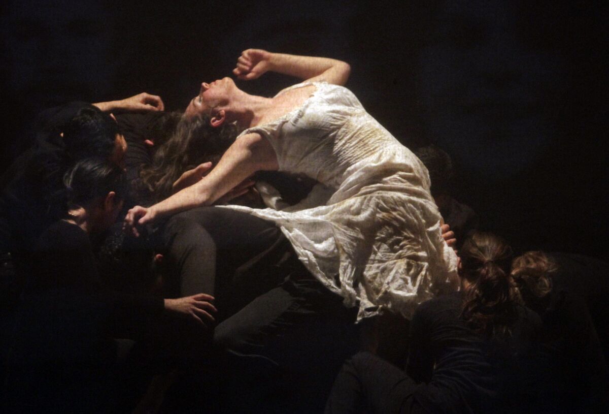 A woman in a white dress leans on and is carried by crouched dancers in dark clothing.
