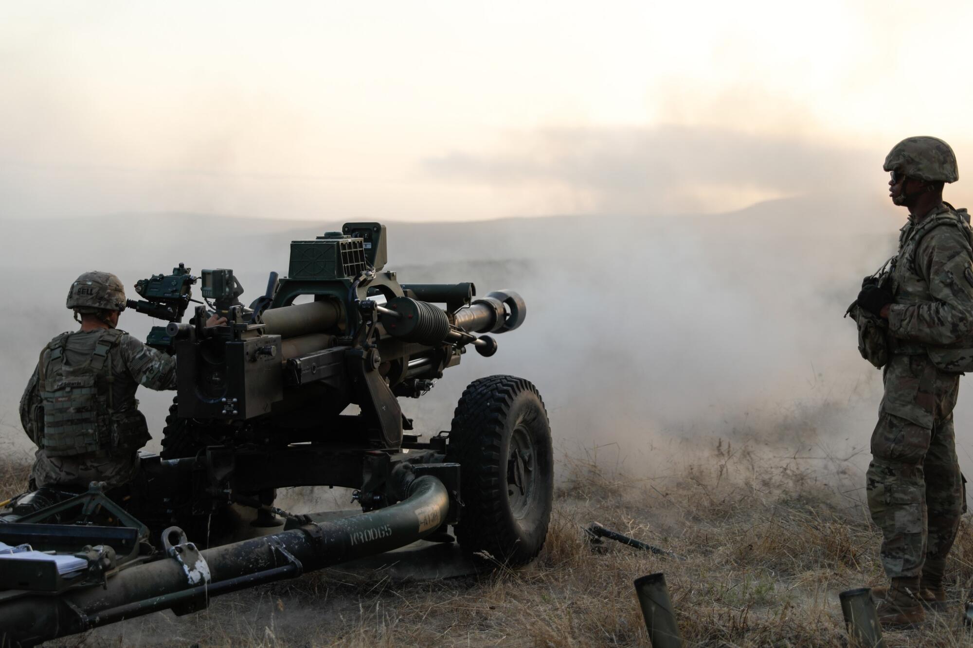 A person in military fatigues fires heavy artillery as another person stands nearby