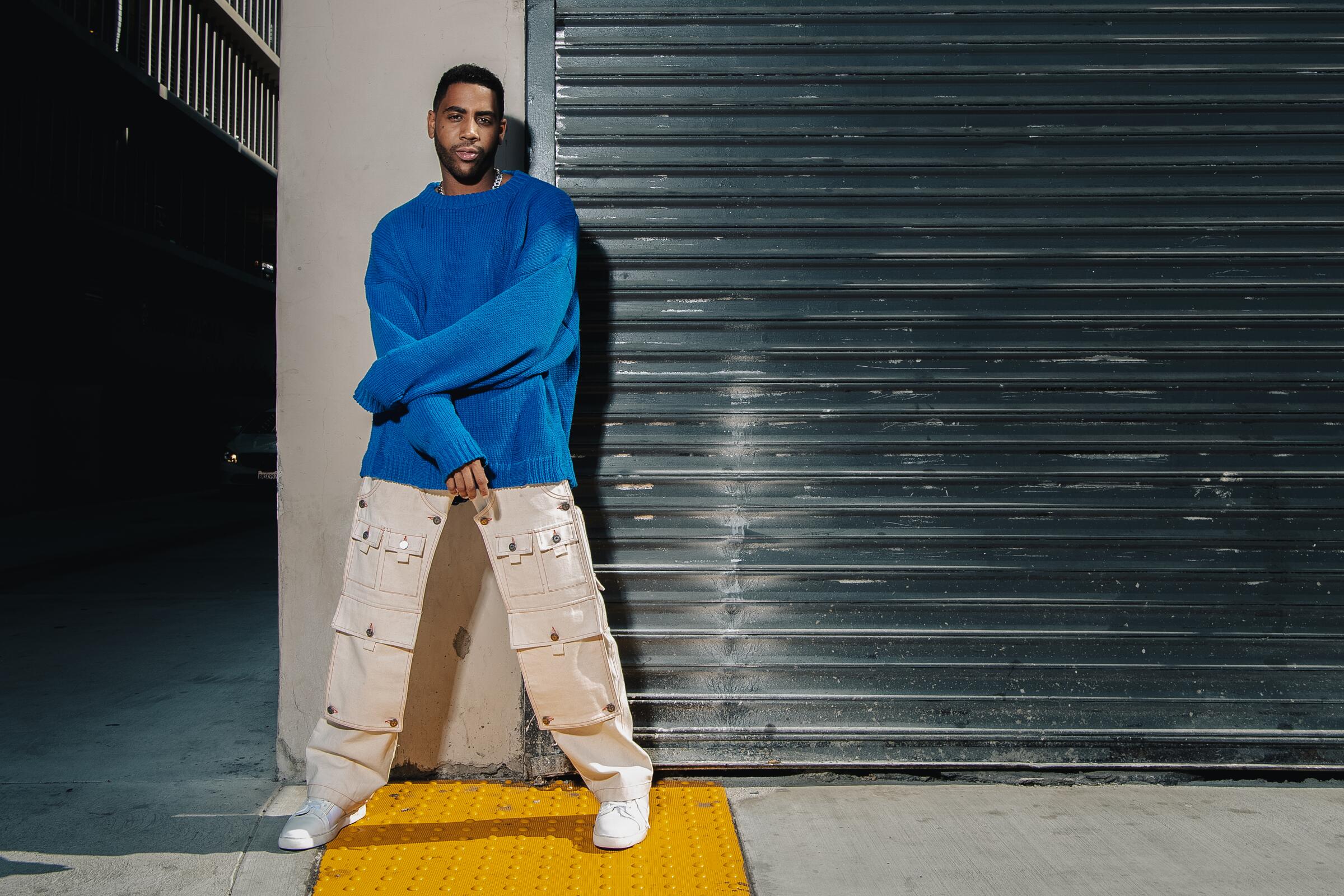 Jharrel Jerome stands near a metal gate in a blue sweater and khaki cargo pants.