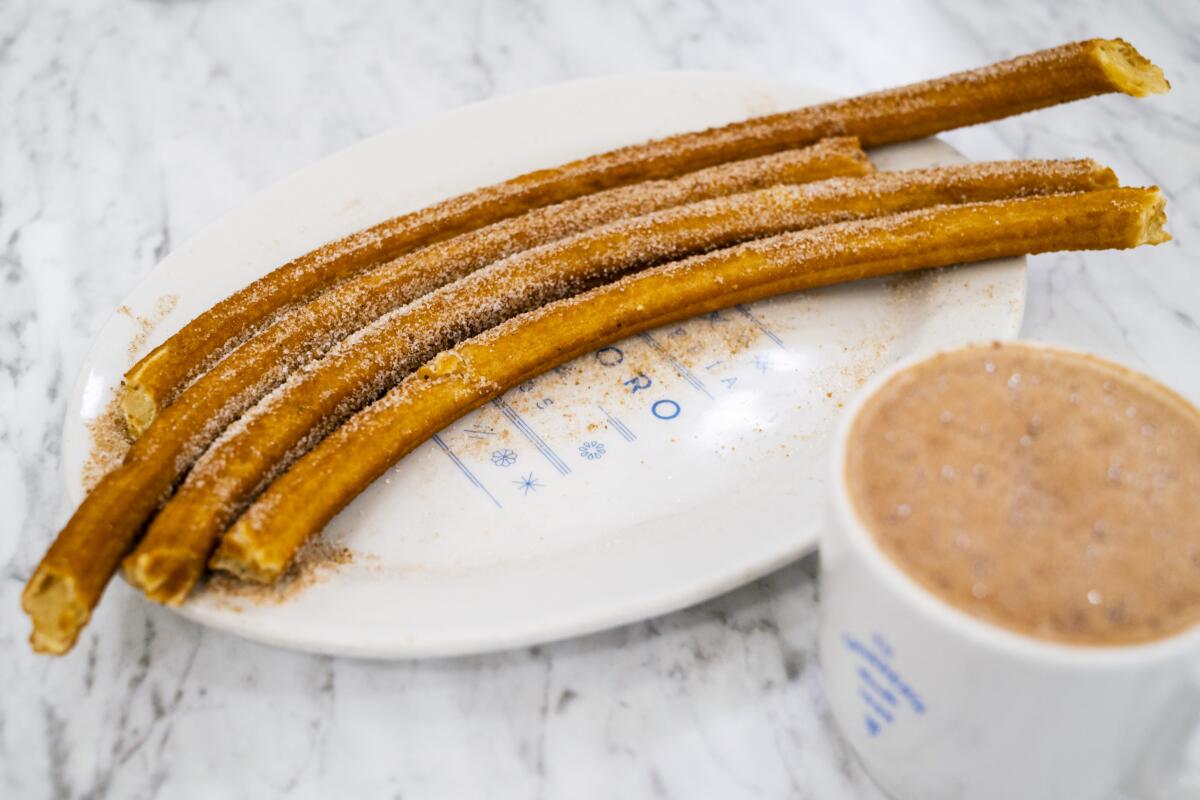 Four churros on a plate next to a cup of hot chocolate.