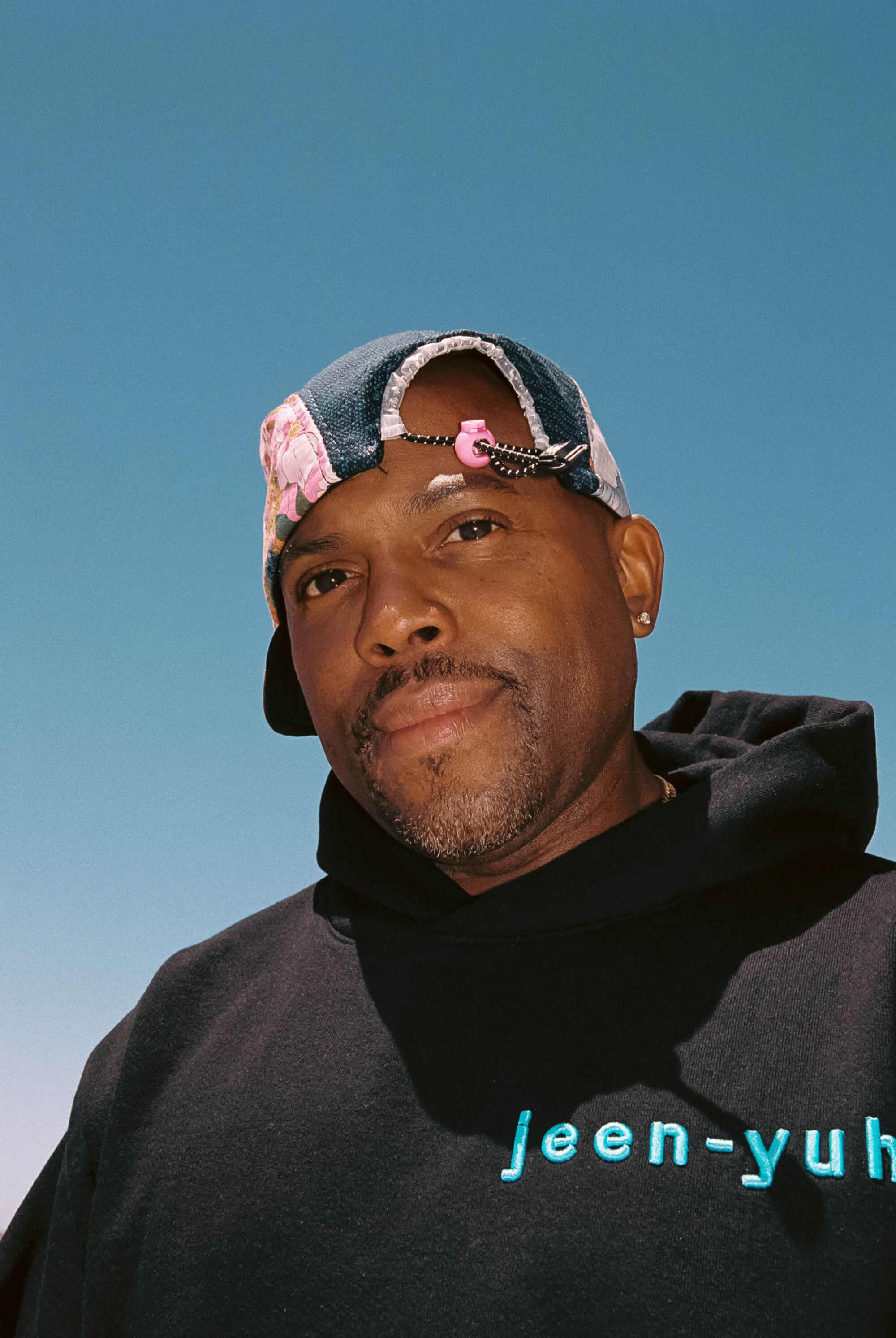 A man poses for a portrait wearring a hat and a sweatshirt, with the sky as a backdrop.