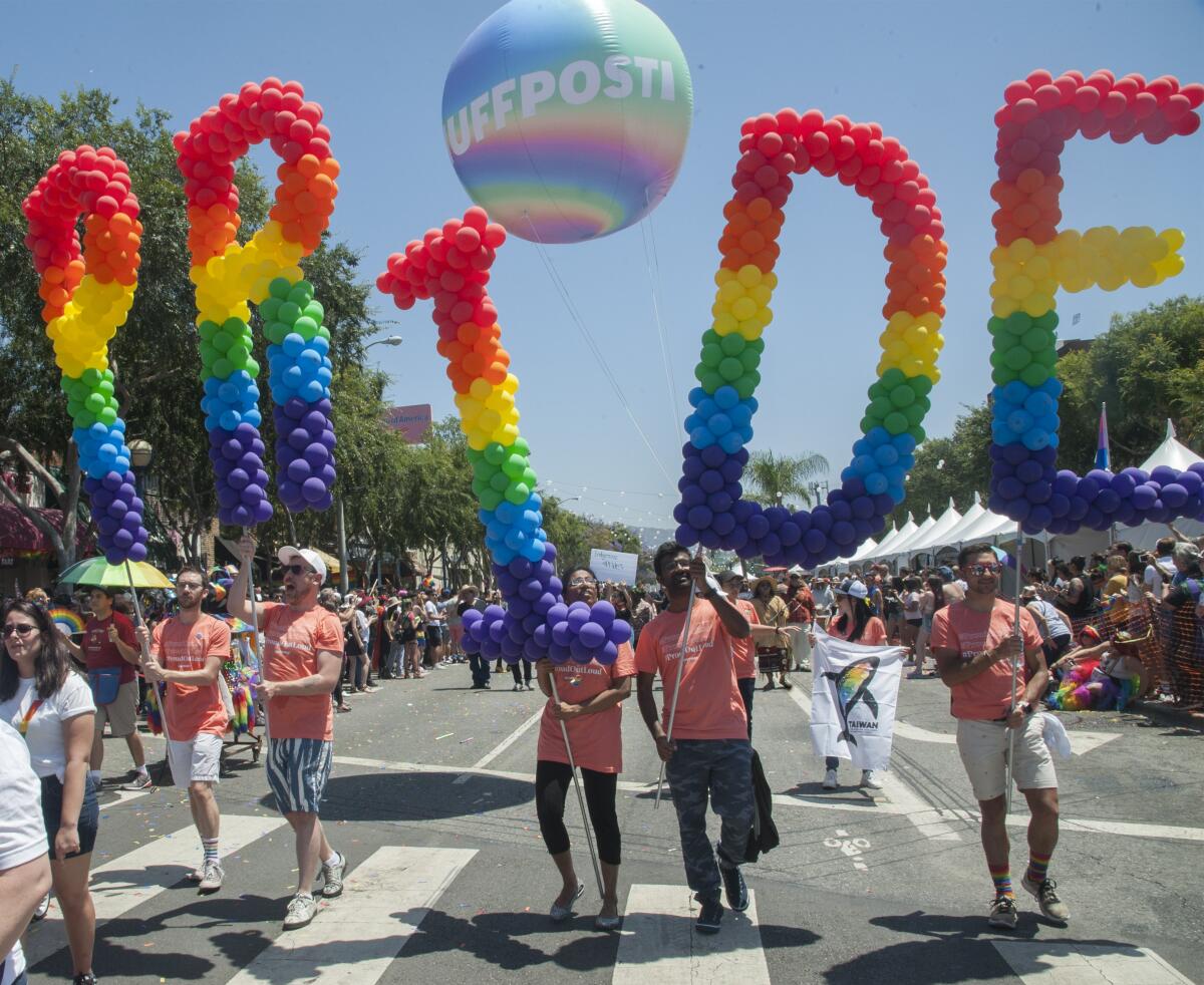 People walk in a parade under rainbow balloon letters spelling the word Pride