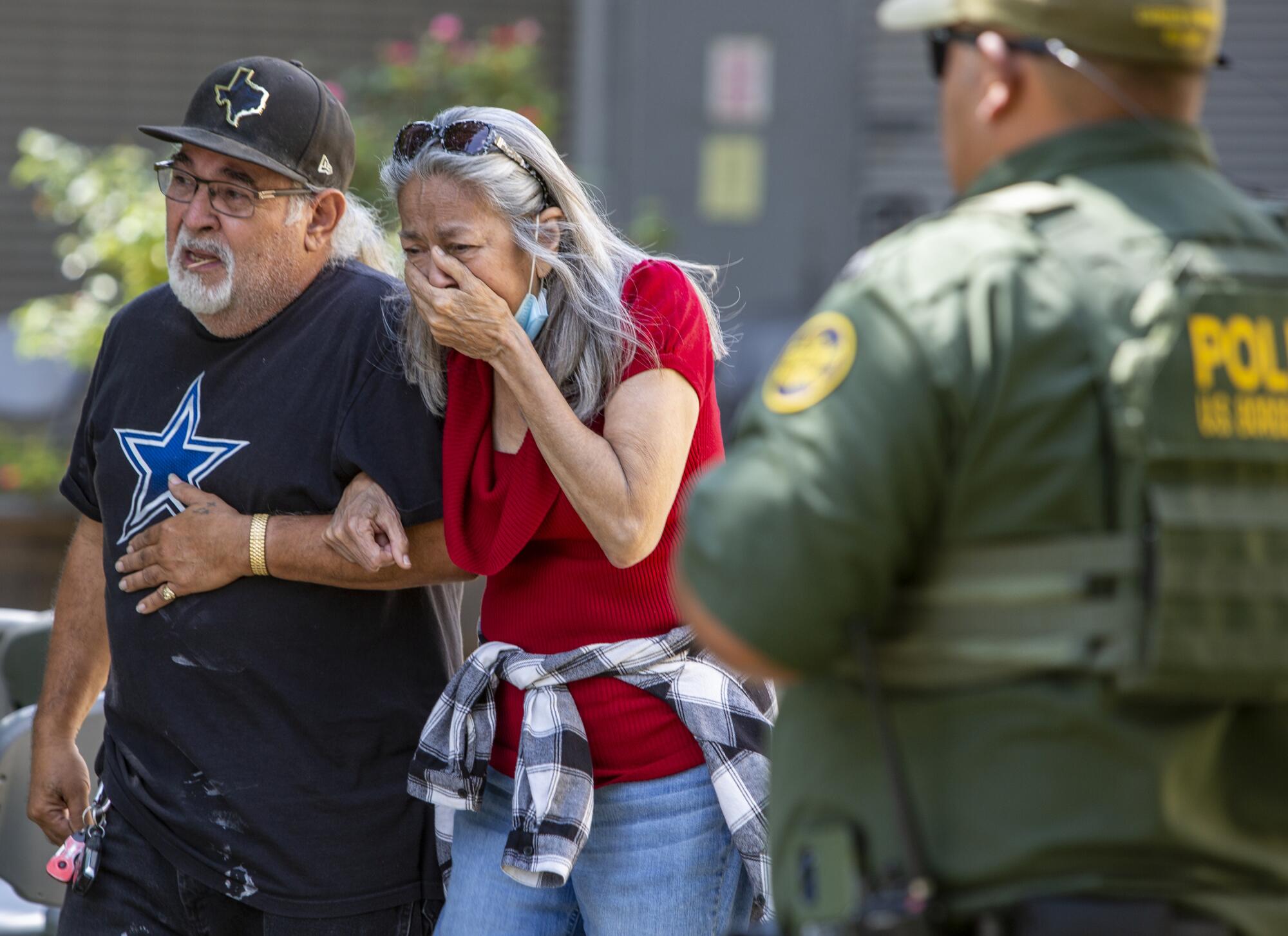 A woman in a red top and jeans cries as she holds the arm of a man in dark clothes 