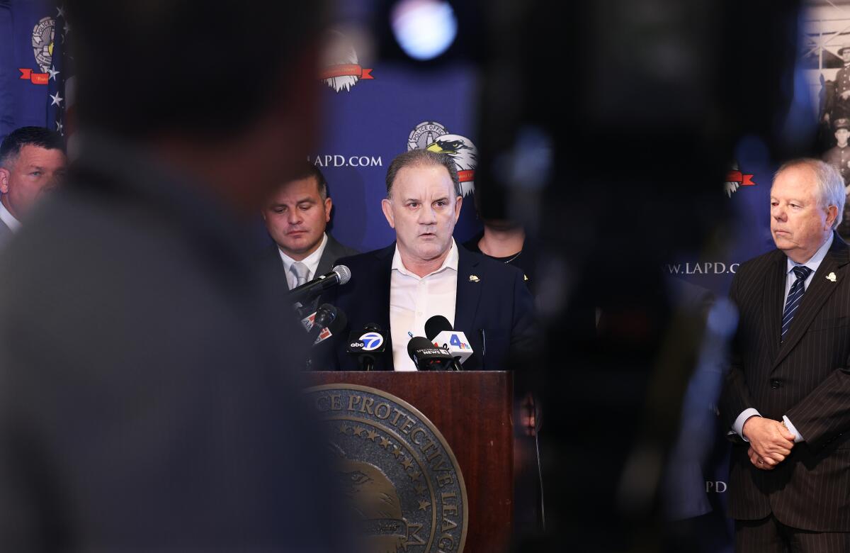 A partially obscured view of Robert Rico speaking from a lectern against a blue backdrop with eagle logos as others stand by