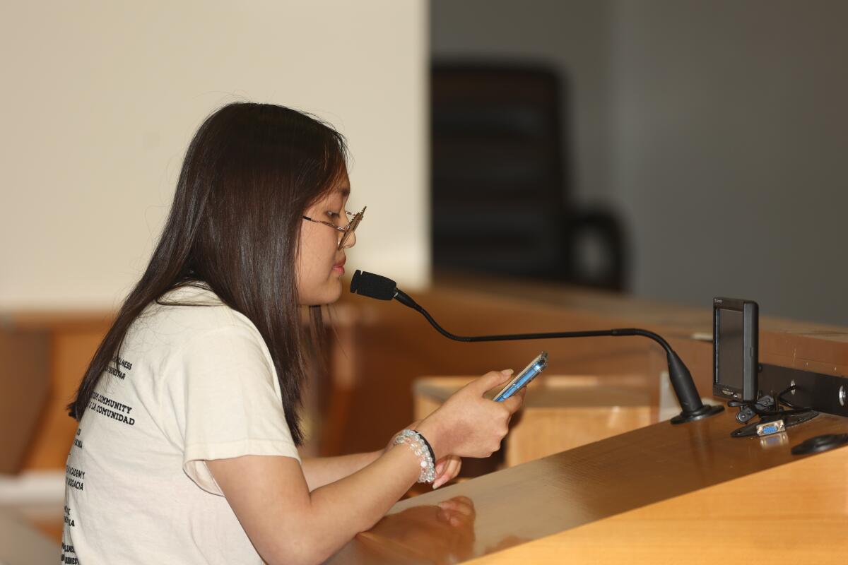 A girl with long hair and glasses looks down at her cellphone while speaking into a microphone 