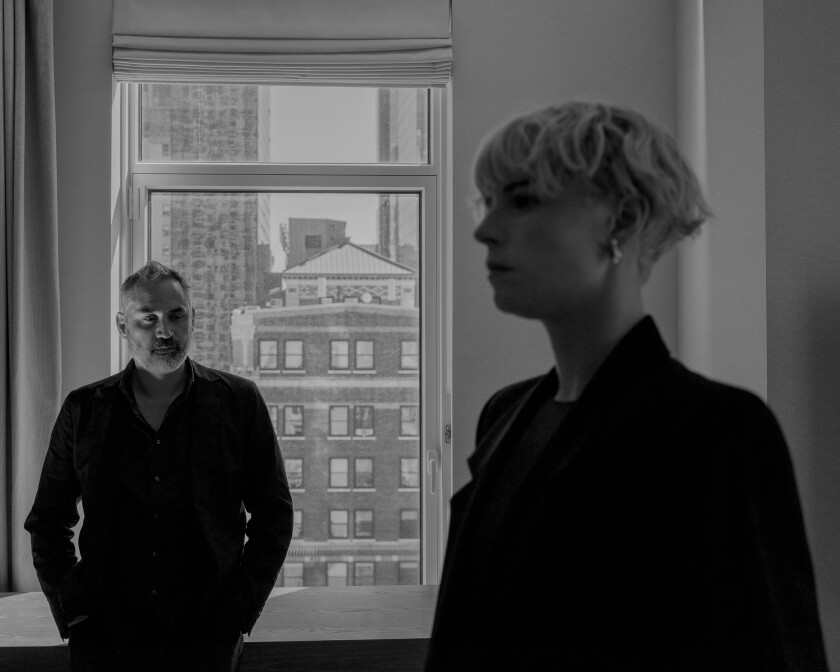 black and white photo of a man, left, and a woman with short hair, tall buildings through a background window