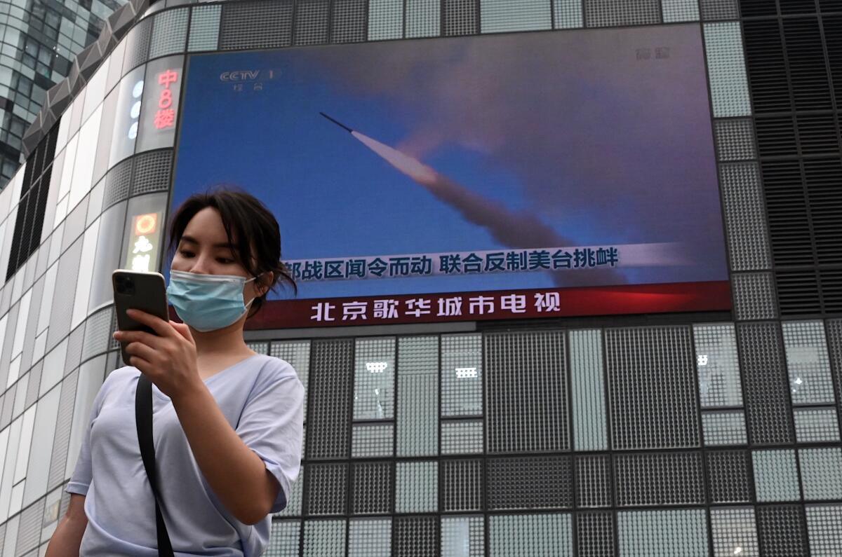 A person using a phone walks in front of a large screen showing a news broadcast about China's military exercises