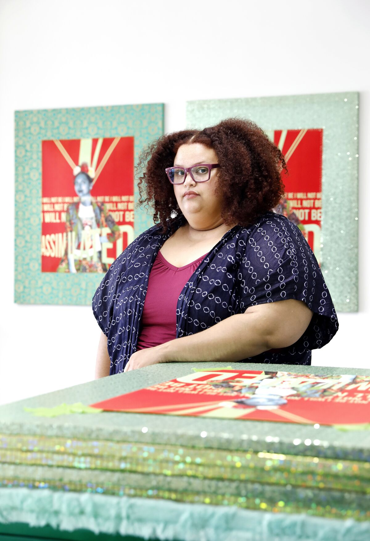 April Bey stands behind a stack of canvases wrapped in shiny green fabrics.