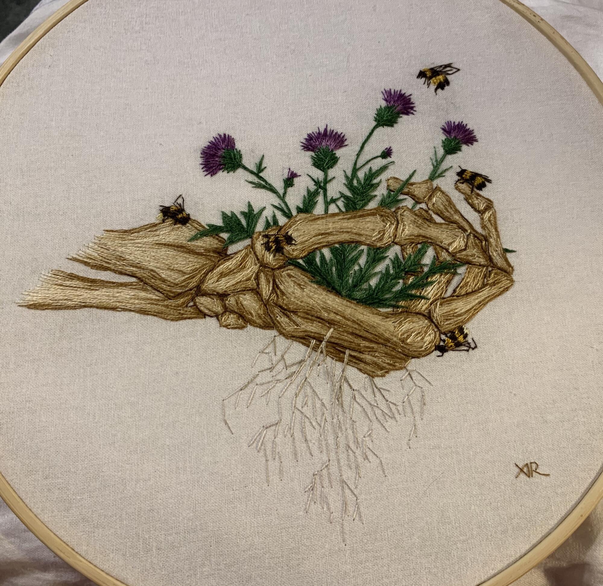 A needlework piece showing a skeletal hand with flowers growing out of it