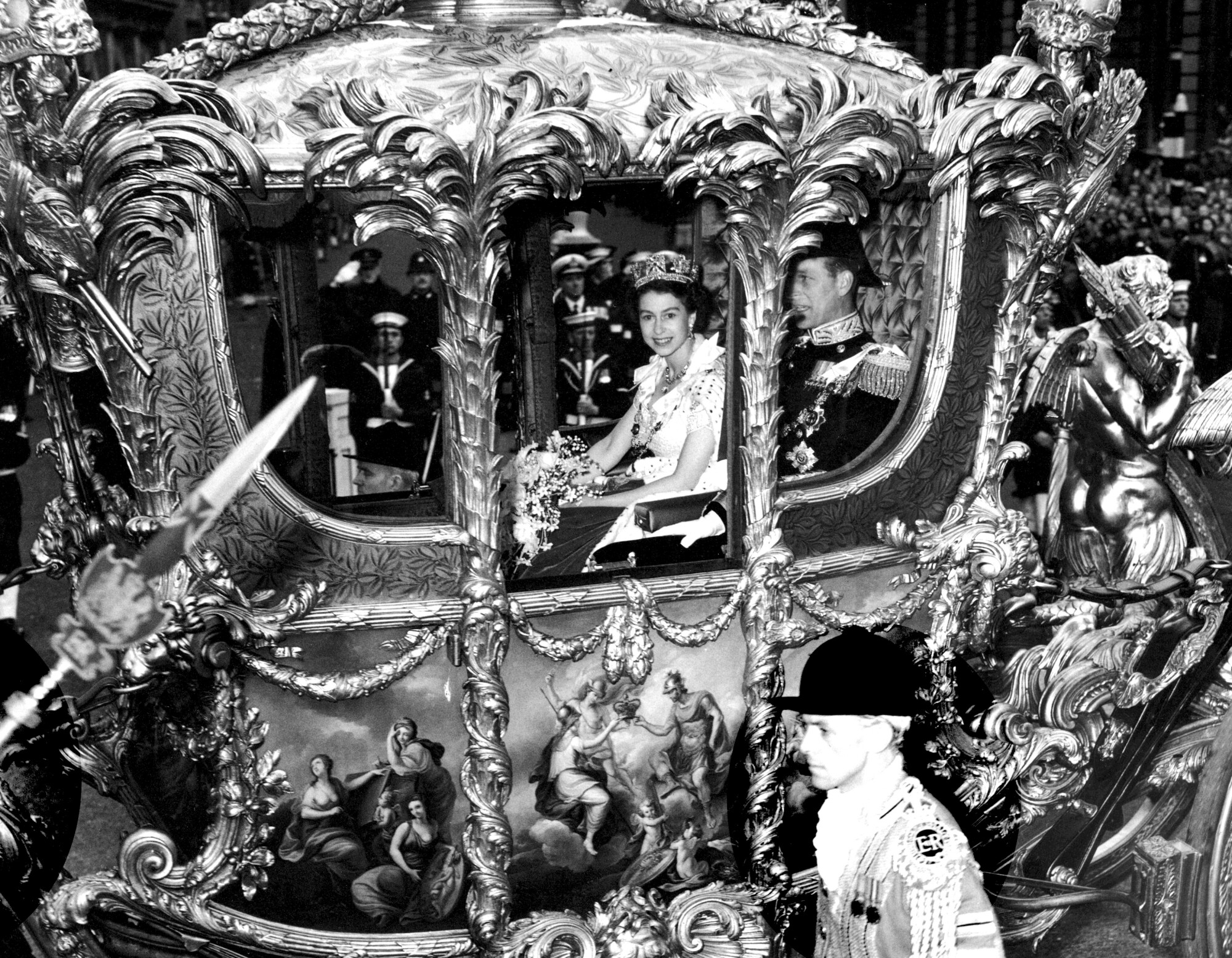 A woman wearing a crown and a white dress smiles as she rides in an ornate carriage beside a man in military uniform 