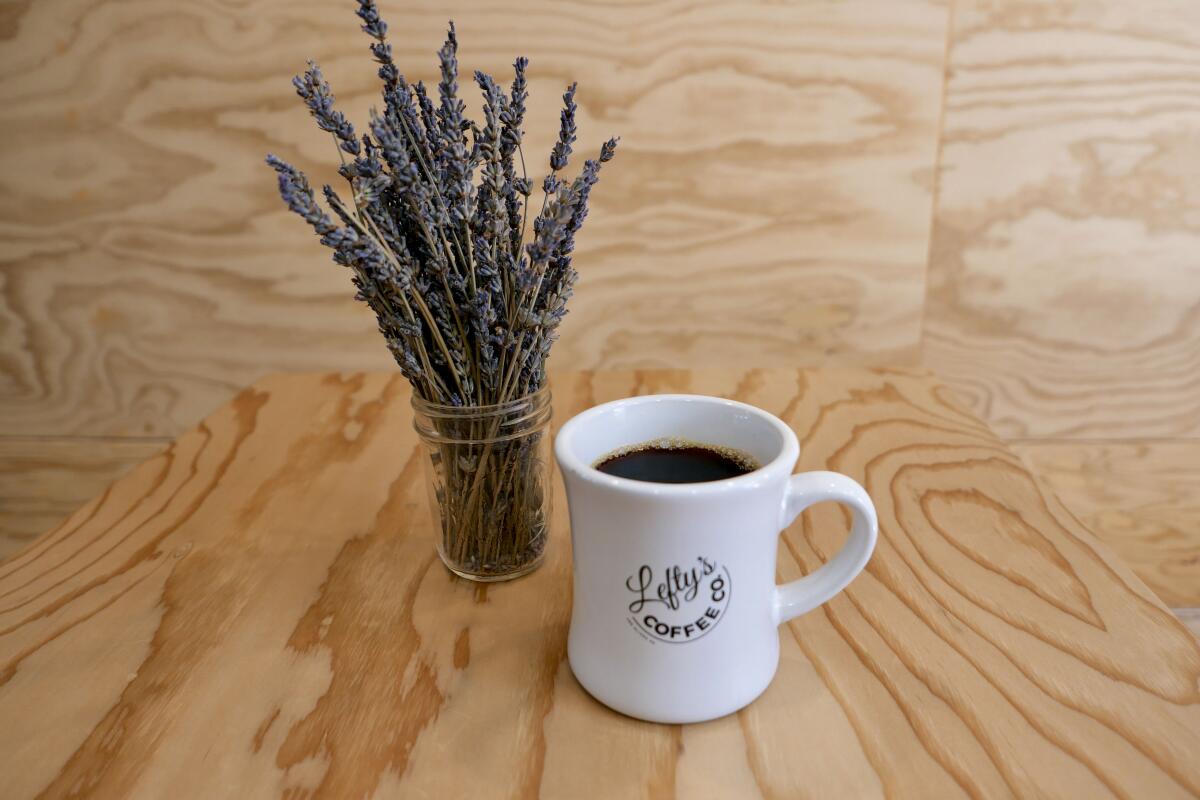 A mug of coffee sits next to a glass jar full of lavender stalks on a wooden table.