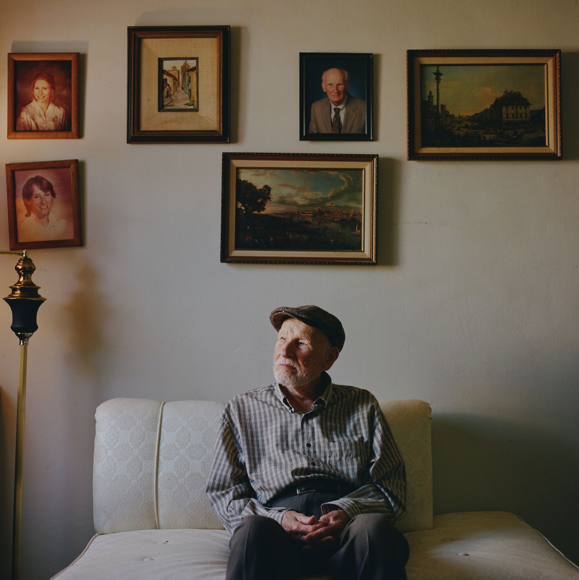 Andrzej Stefanski looks out of the window in his living room. Above him hang paintings of Warsaw and photos of his family.