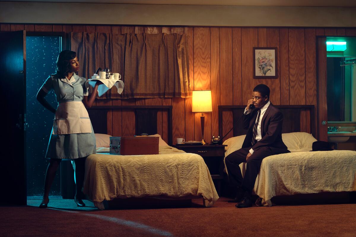 A man sits on a hotel room bed, talking on the phone, when a maid walks in holding a room service tray