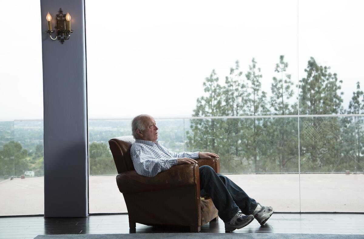 Seymour Stein sits in a chair in front of a glass wall overlooking the outdoors.