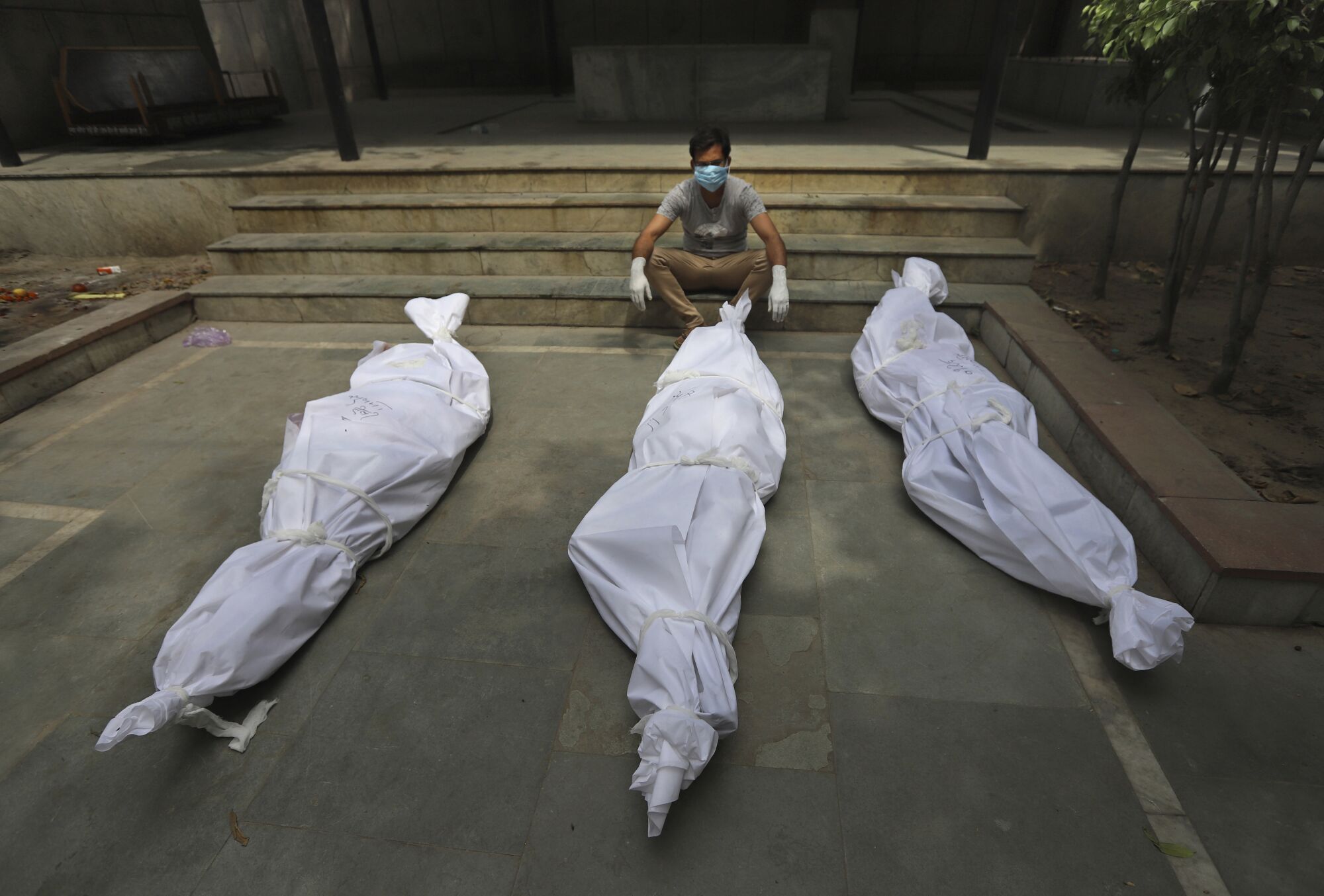 A man sits on steps. Before him are three bodies shrouded in white. 