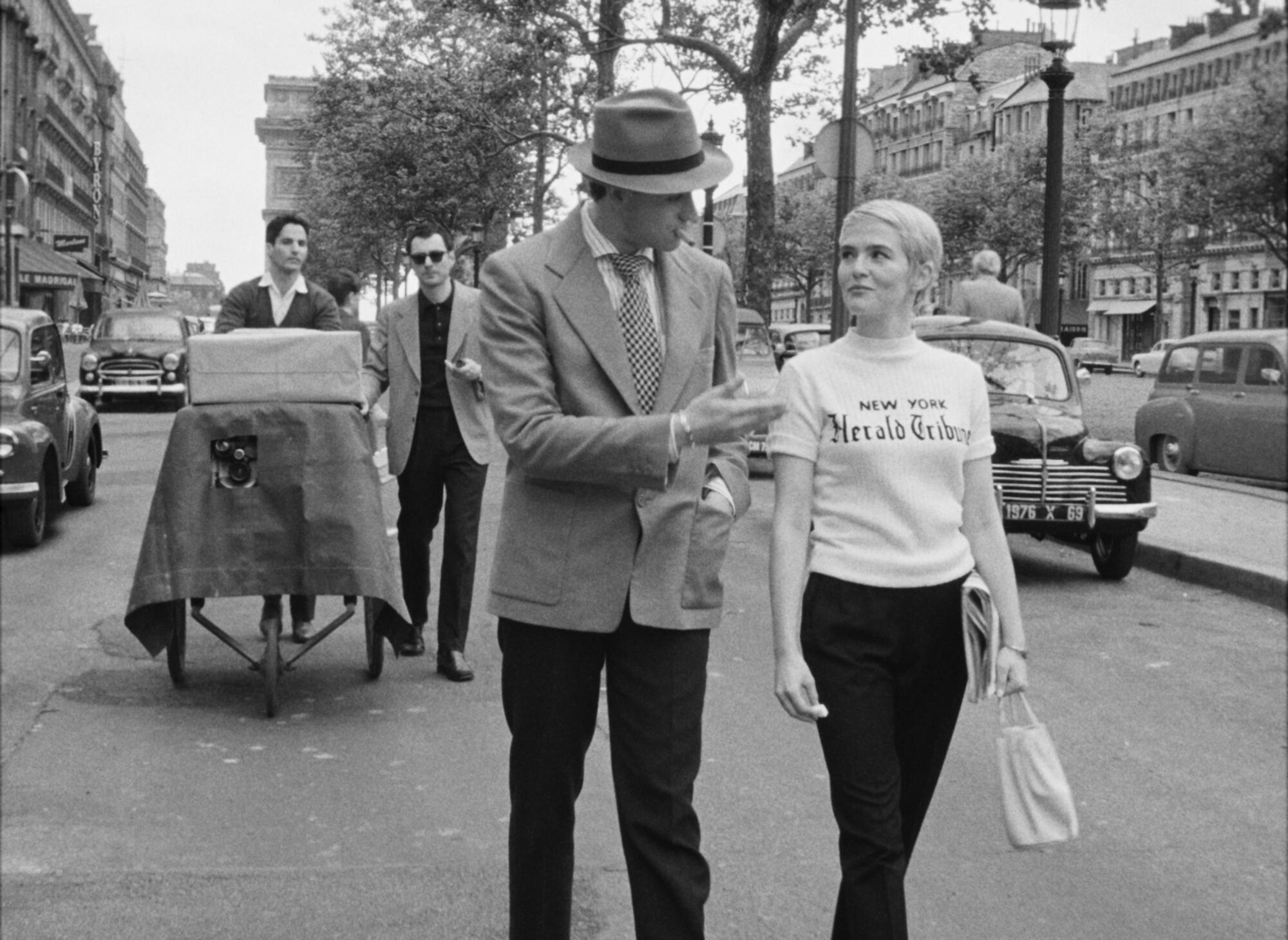 A man in a fedora strolls with a woman wearing a white T-shirt while two men photograph them from behind.