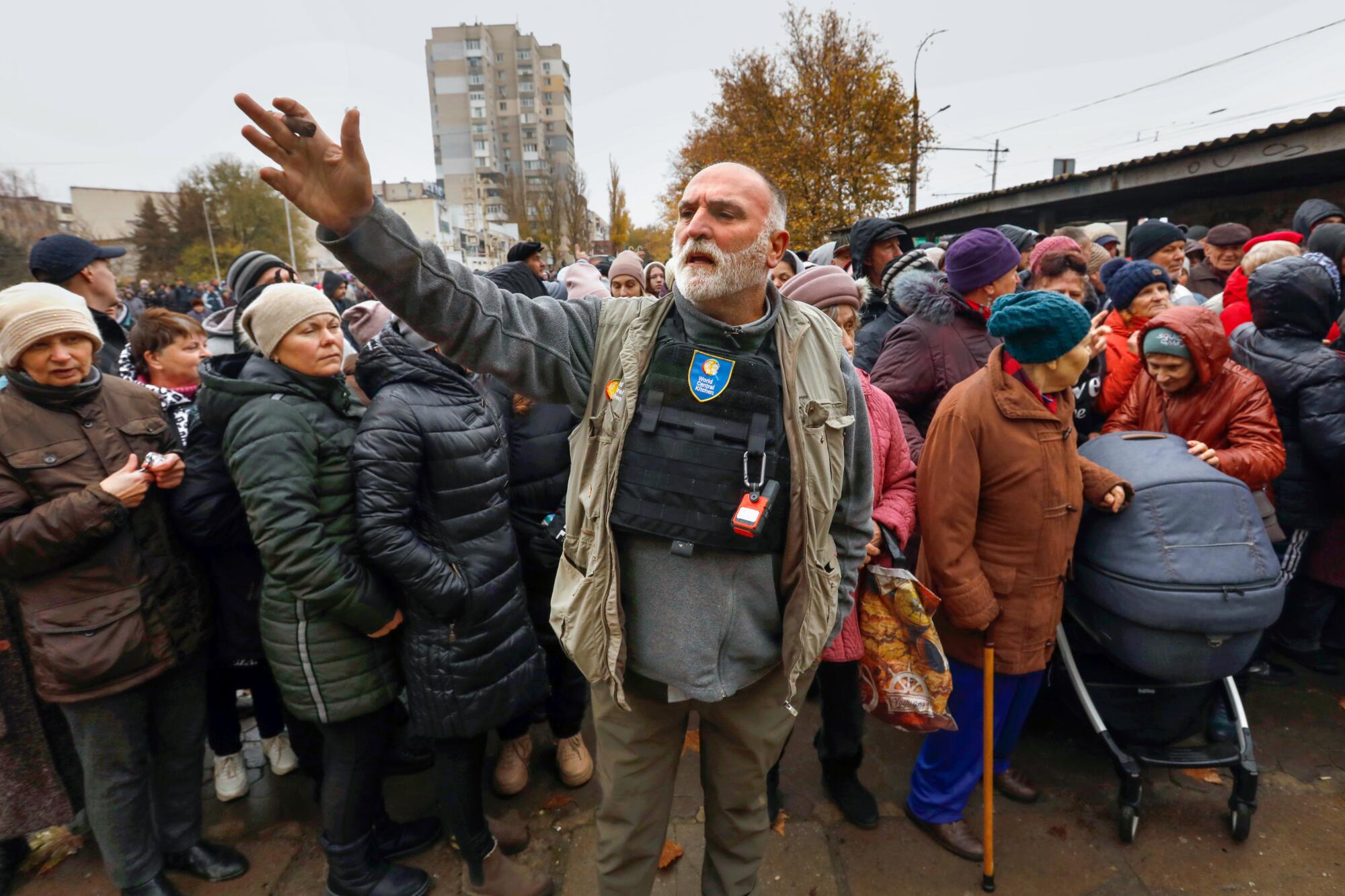 A man with a gray beard, in a khaki vest, has one hand outstretched. Behind him is a large crowd of people 