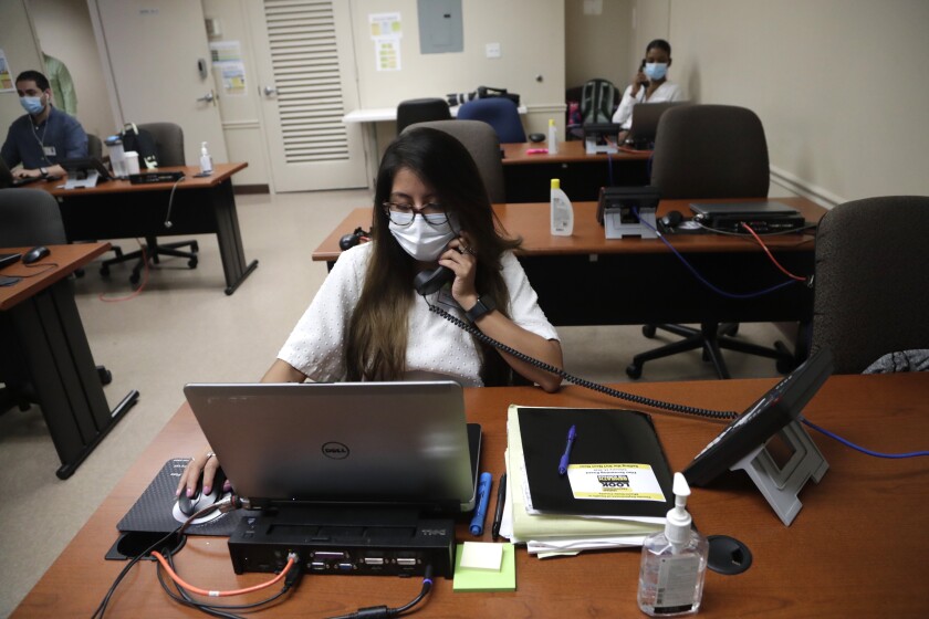 A woman in a face mask sits at a desk speaking into a phone as she looks at a laptop.