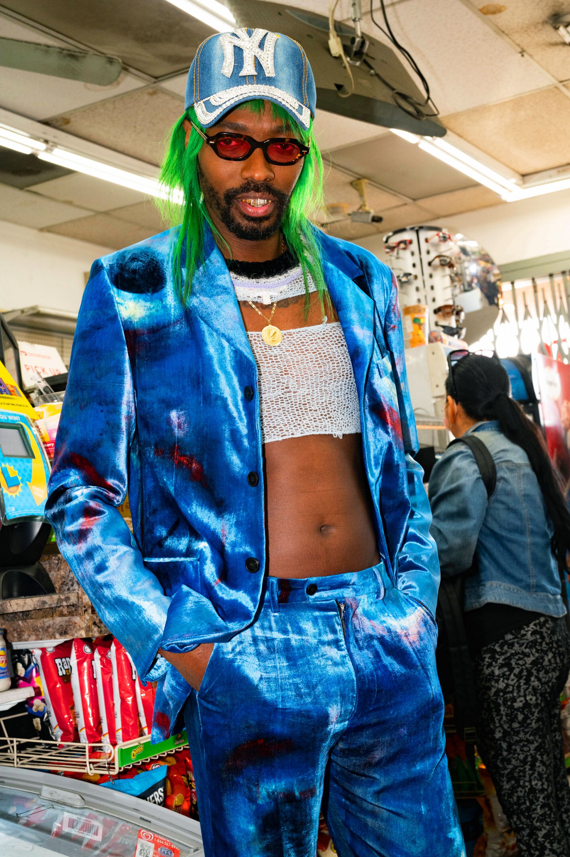 A person wearing a blue suit, crochet crop top and blue cap stands in a convenience store.