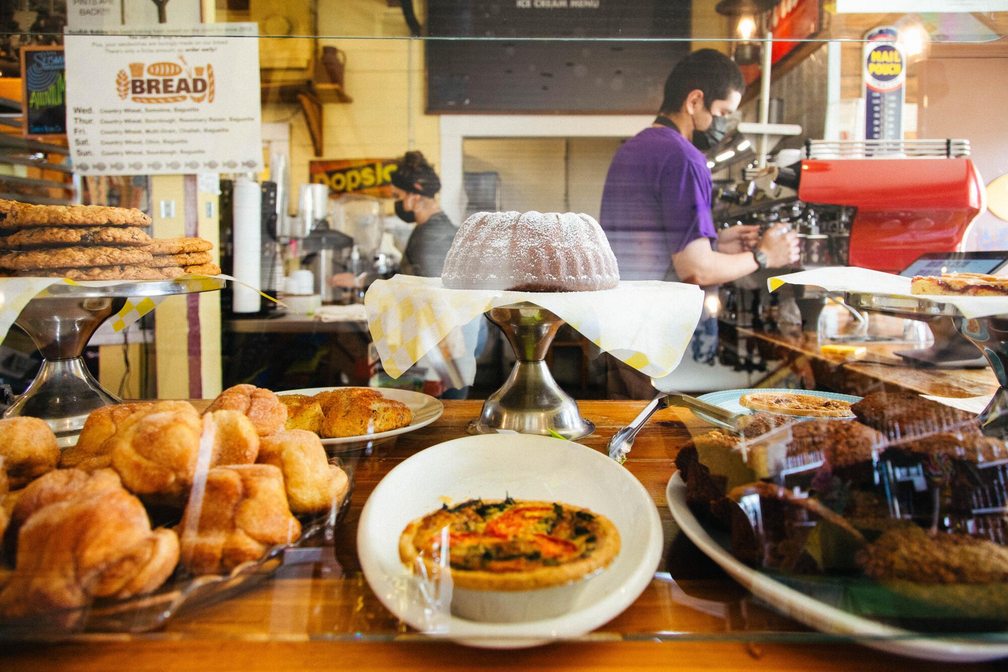 A counter of baked goods behind glass, with a person making coffee  