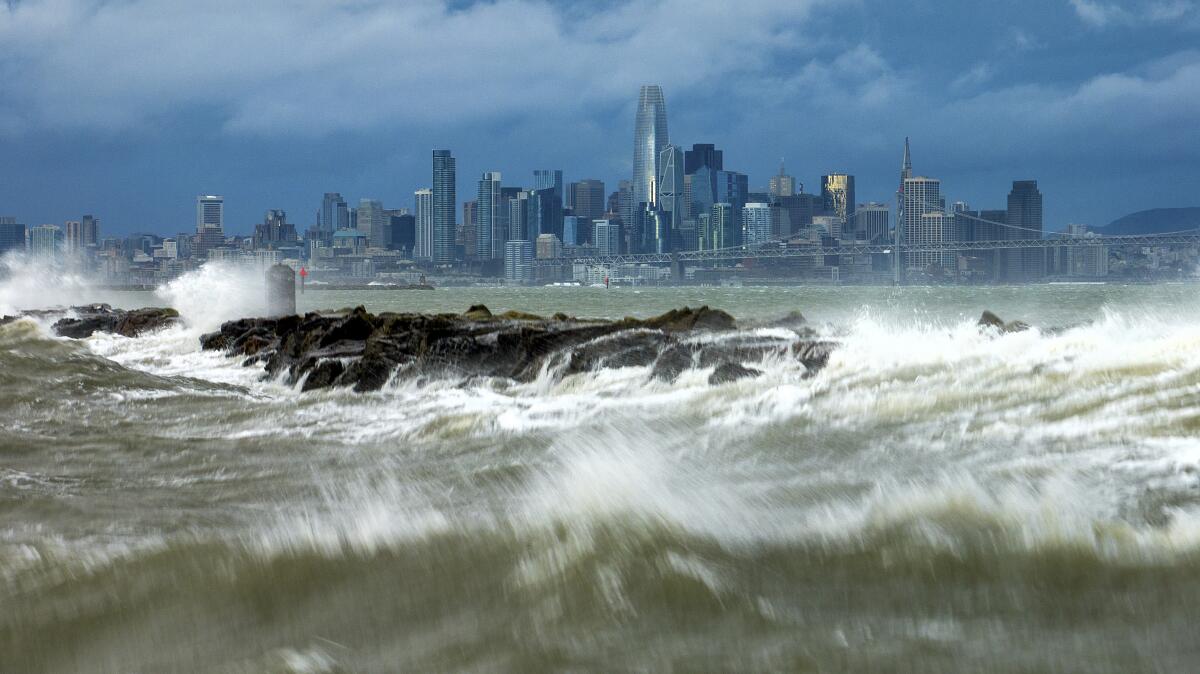 Closeup of rocks and white-capped surf with a city skyline in the background.