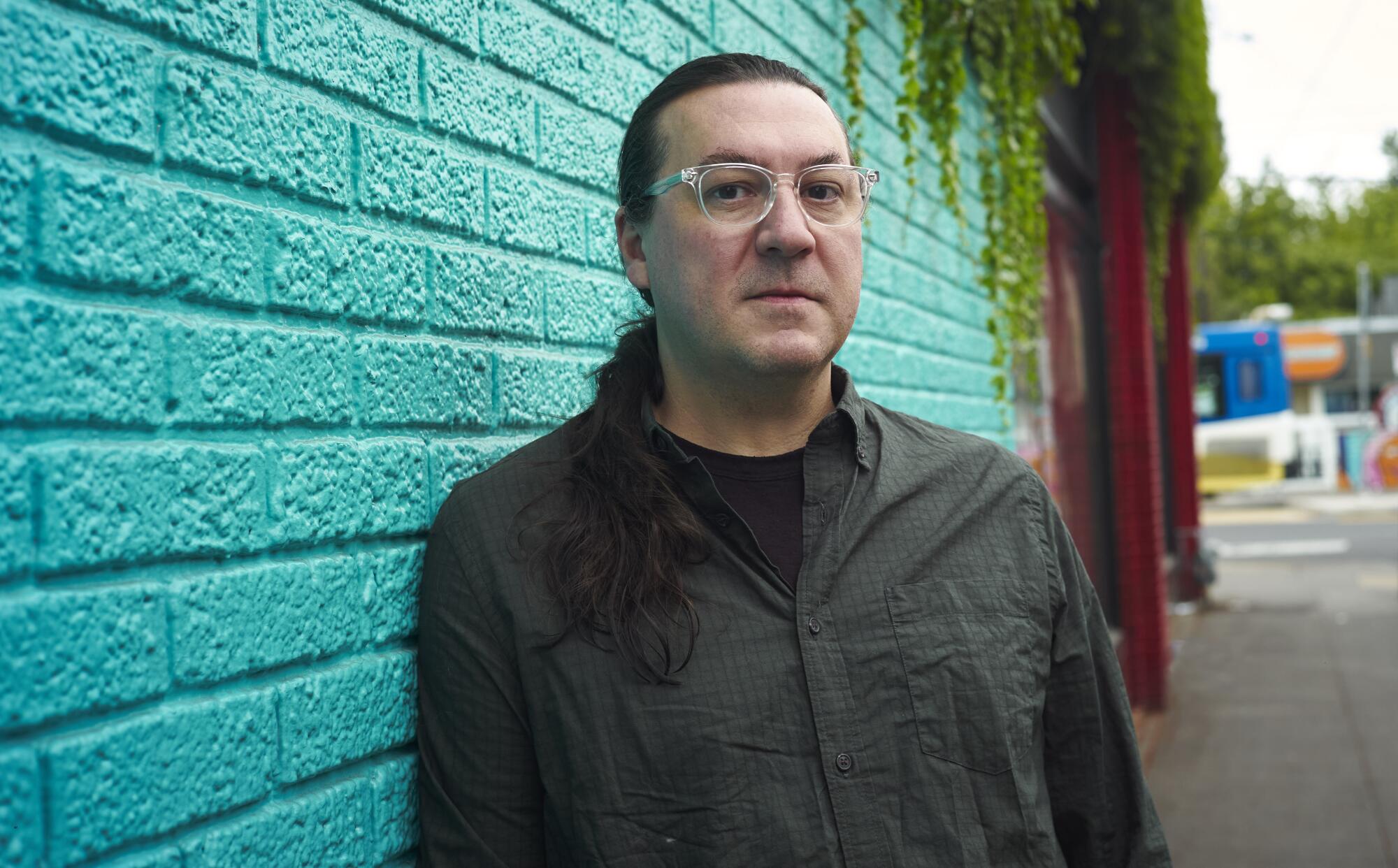 A man with wire-rimmed glasses and a ponytail poses outside against a turquoise wall. 