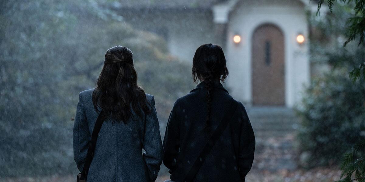 Two people are seen from behind as they stand in front of a house amid precipitation.