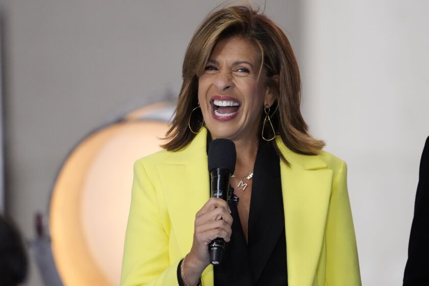 A woman with short brown hair smiling while holding a microphone and wearing a bright yellow coat