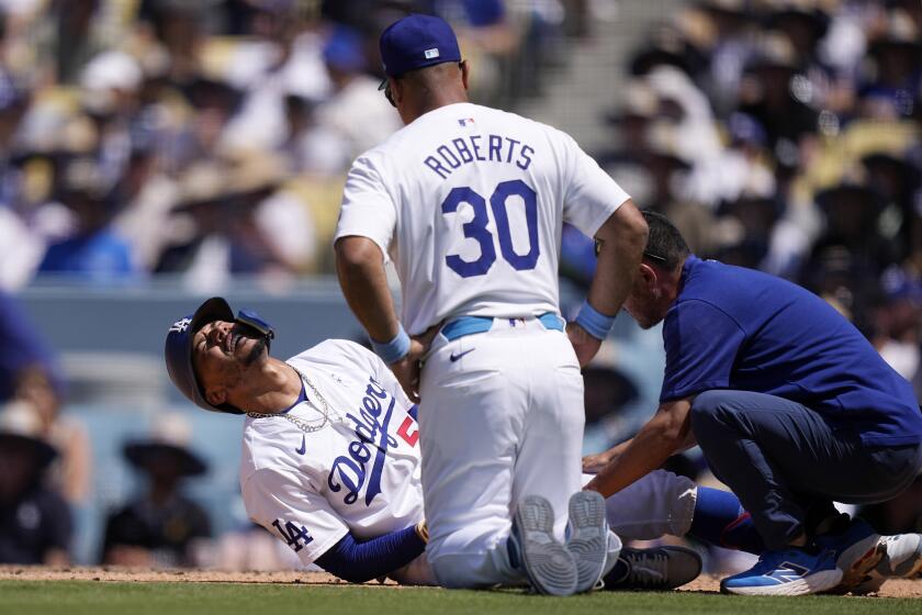 Mookie Betts, de los Dodgers de Los Ángeles (izquierda), se retuerce de dolor sobre el terreno tras ser golpeado por un lanzamiento, mientras que el mánager Dave Roberts y un entrenador lo asisten durante la séptima entrada del juego de béisbol ante los Reales de Kansas City, el 16 de junio de 2024, en Los Ángeles. (AP Foto/Mark J. Terrill)