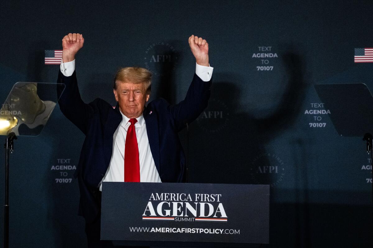 Former President Trump holds up two fists at a lectern reading "America First Agenda."