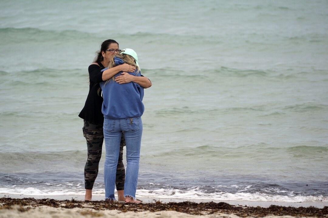 On the sand, with waves lapping in the background, a woman holds another woman, who leans into her shoulder.